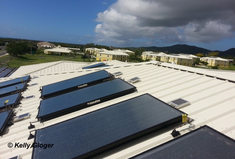 Multiple flat-plate solar collectors on white metal roof with housing complex view