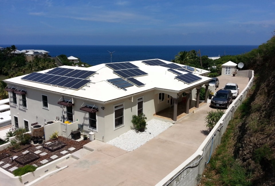 Aerial view of large residential home with extensive solar PV array on white flat roof overlooking Caribbean sea