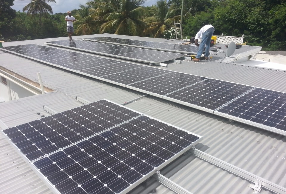 Two installers working on solar PV panels on corrugated metal roof with palm trees in background